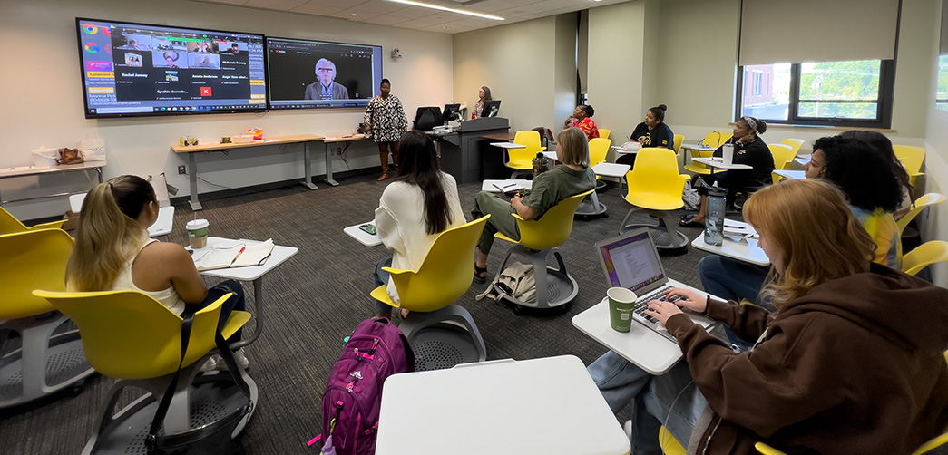 Students are seated in gold chairs in a classroom while an instructor speaks. Students joining virtually are displayed in the background on a screen.