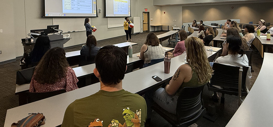 A group of students are seated as two presenters stand at the front of a room, holding colored beachballs.
