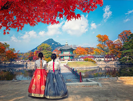 Two people in traditional dresses stand in the foreground with bright red foliage above them, staring at a pagoda and mountaintop in the distance.
