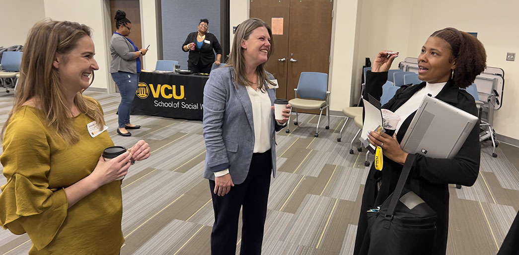 Three people stand talking and laughing. Table cover, at rear, reads VCU School of Social Work.