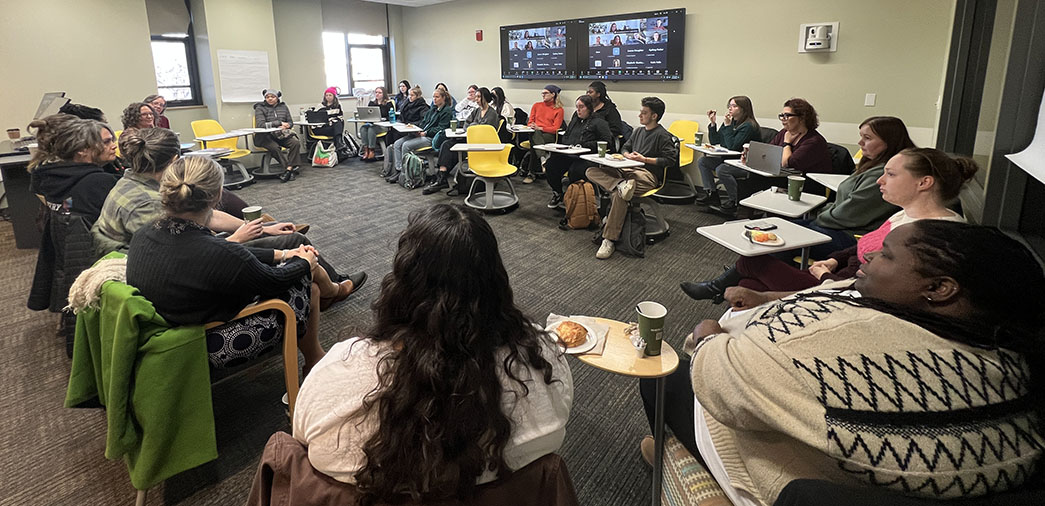 A large group of students, alumni and faculty sit in a circle with additional participants joining online via digital displays in the back of the room.